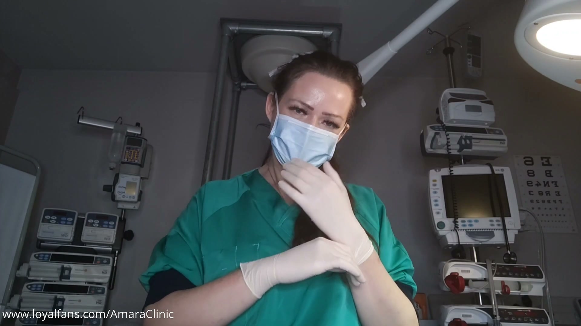Mistress Katharina Amara in green scrubs and surgical mask drawing on gloves during this free Littmann stethoscope fetish video from Amara Fetish Clinic Belgium